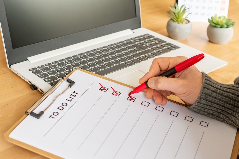 Hand with red marker checking off completed tasks on a to do list clipboard, symbolizing productivity, organization, planning, and goal achievement on a wooden desk with a laptop
