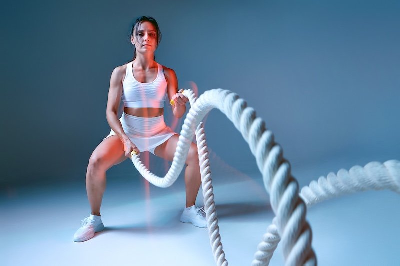A dynamic scene of an energetic woman engaging in a workout with battle ropes in a studio environment. She is dressed in a white sports outfit and sneakers, showcasing focused determination.